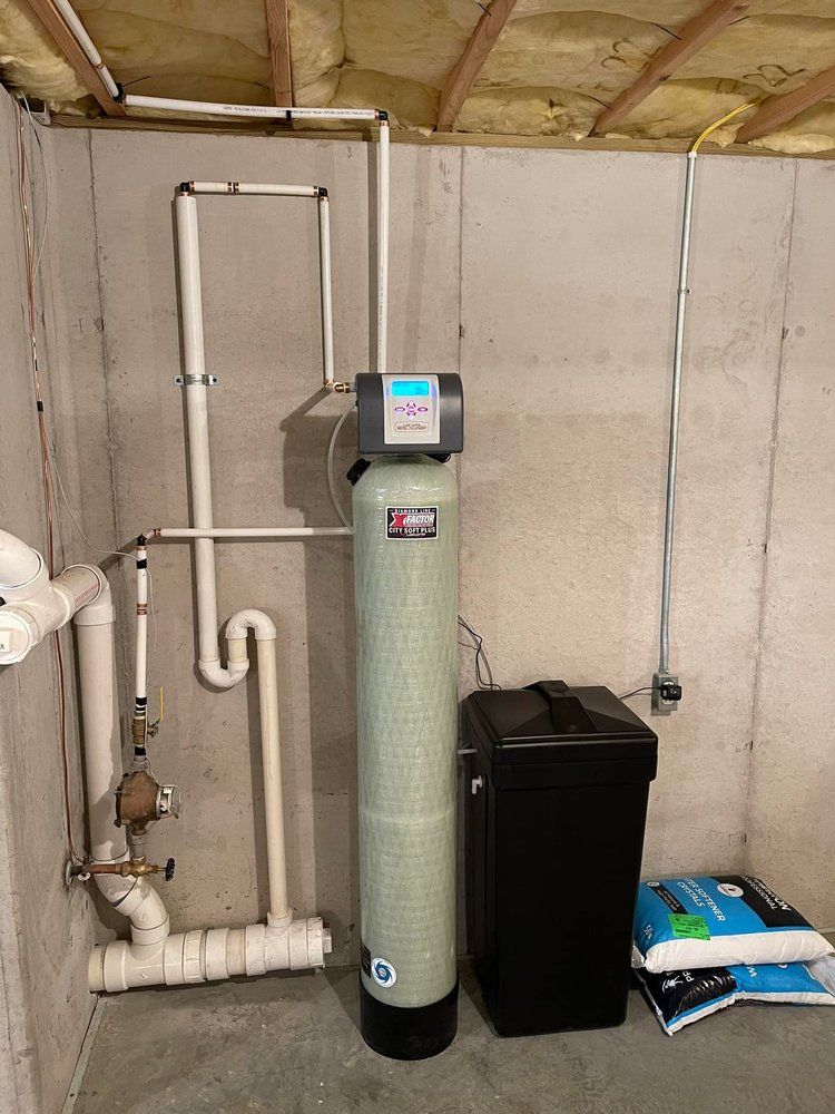 A water softener is sitting in a basement next to a black trash can.