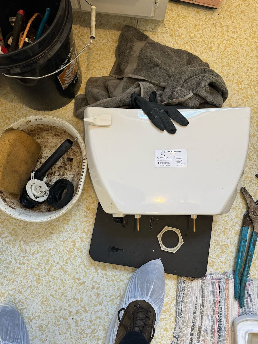 A toilet tank is sitting on the floor next to a bucket and a pair of pliers.