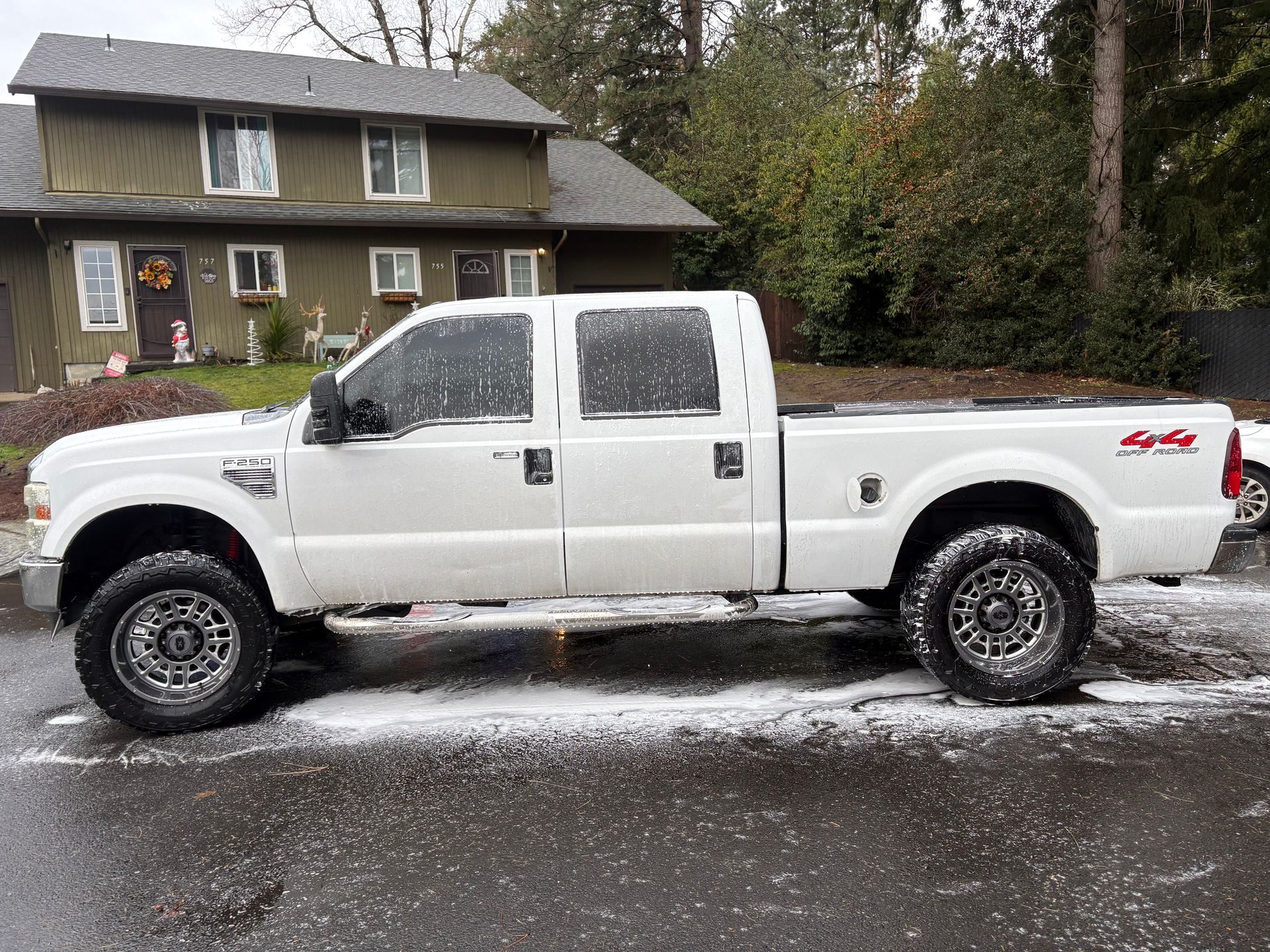 A white Ford F-250 crew cab pickup truck parked on a wet, paved driveway in front of a green two-story house.