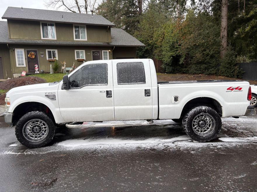 A white Ford F-250 crew cab pickup truck parked on a wet, paved driveway in front of a green two-story house.