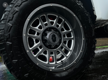A close-up shot of a dark gray, multi-spoke off-road truck wheel with a deep dish rim and a knobby all-terrain tire.