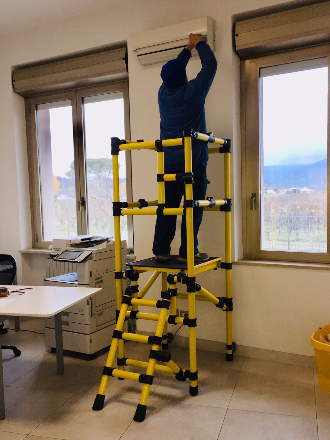 A man is standing on a yellow ladder working on an air conditioner