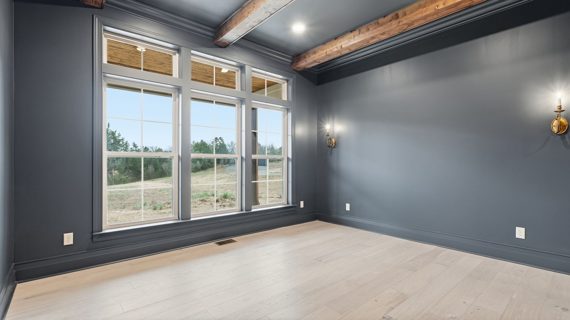 Empty room with large windows, blue-gray walls, wood beams, and light-colored floors.