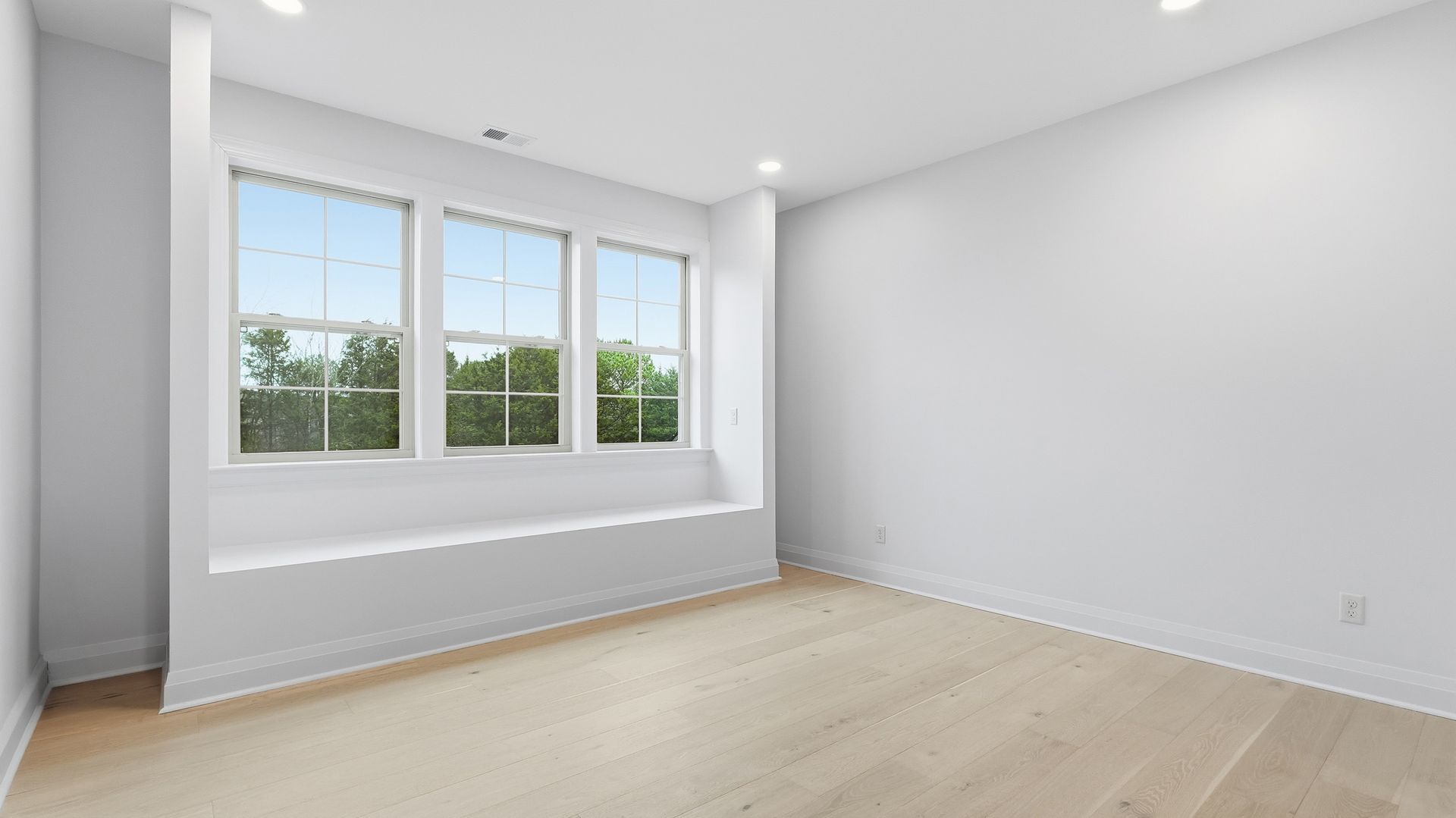 Empty white room with three windows, a built-in bench, and light wood flooring.