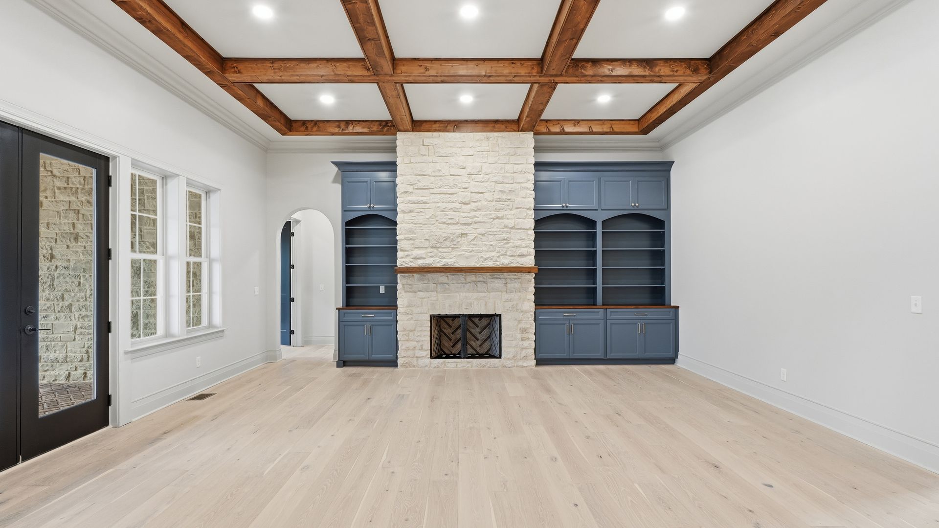 Empty room with a fireplace, built-in shelves, and a wooden beam ceiling. Light wood floors and blue cabinetry.
