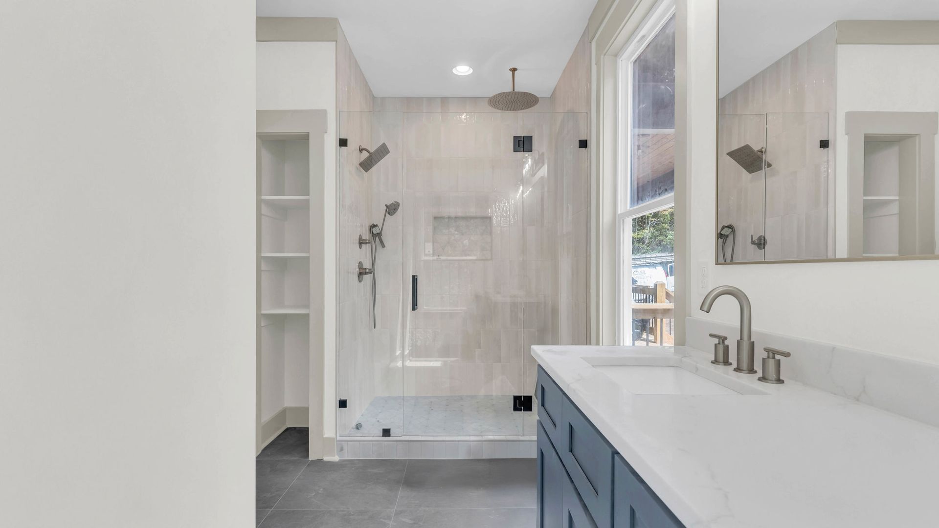 Bathroom with blue vanity, glass shower, and built-in shelving. Light gray tiled walls.