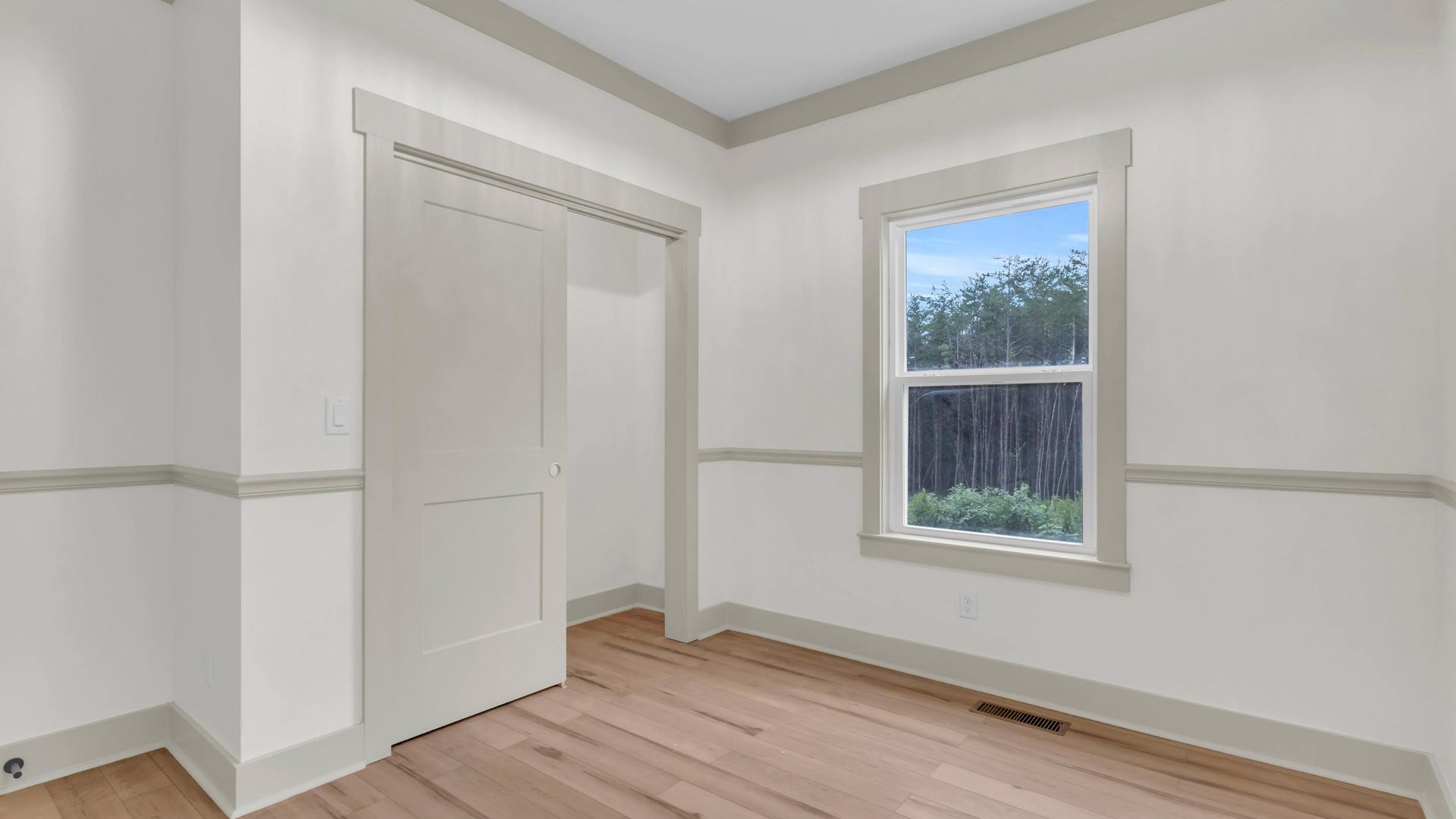 Empty bedroom with wooden floor, closet, and window showing a forest. Light grey walls and trim.