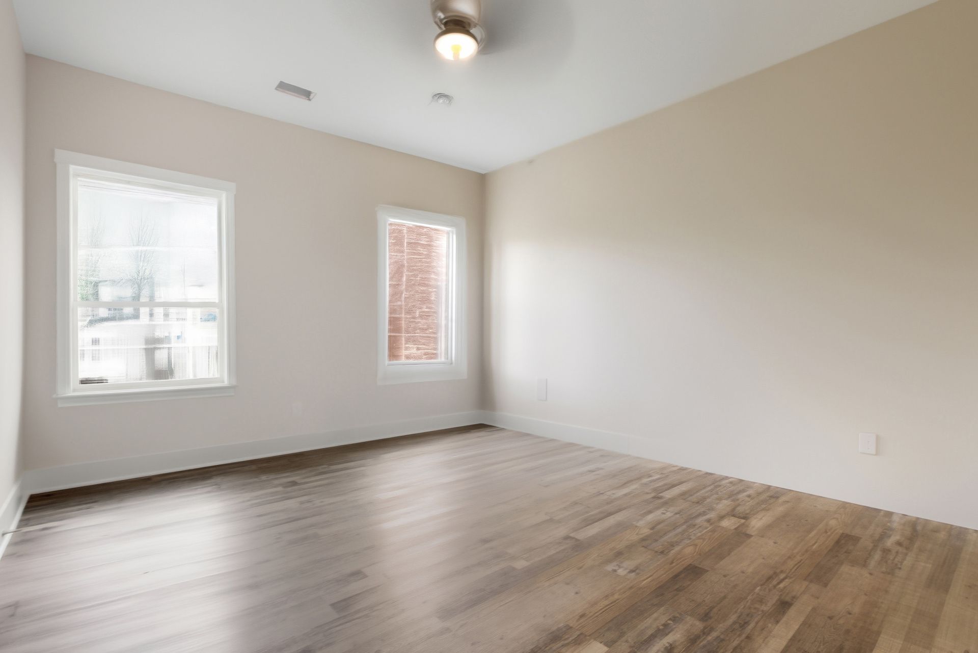 Empty room with hardwood floors, two windows, and neutral walls. Ceiling fan.