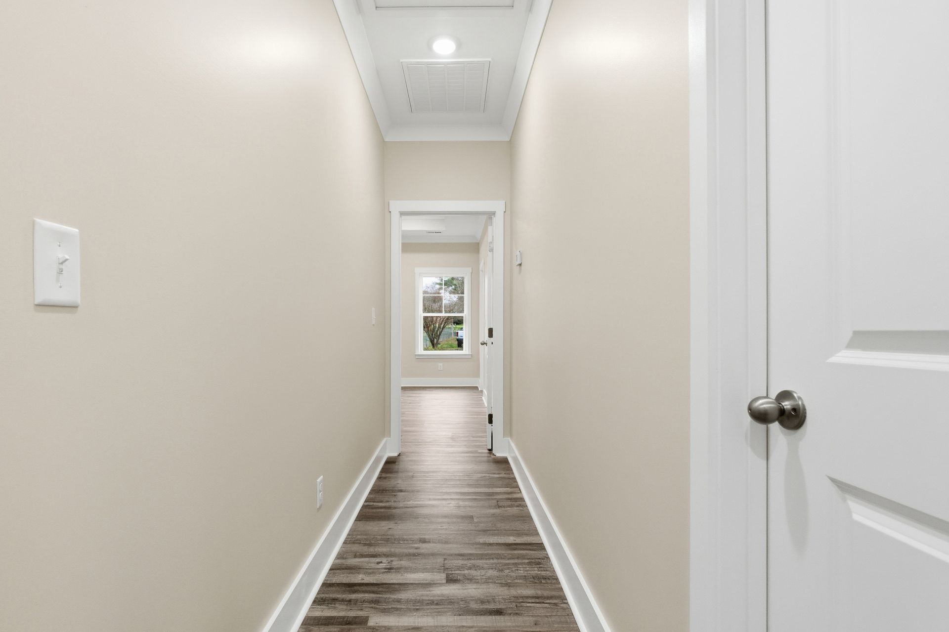 Narrow hallway with light beige walls, wood-look flooring, and a window at the end.