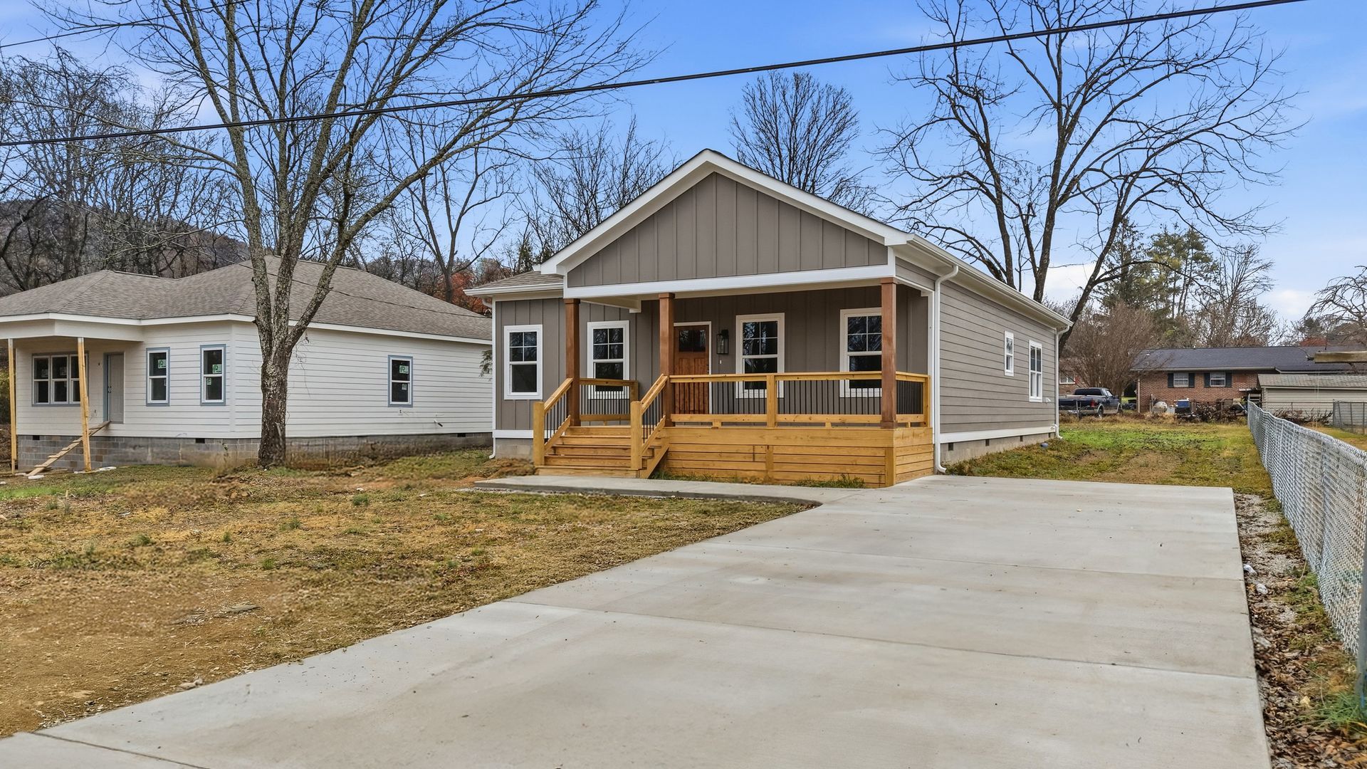 A gray house with a wooden porch and driveway, trees, and another house in the background.
