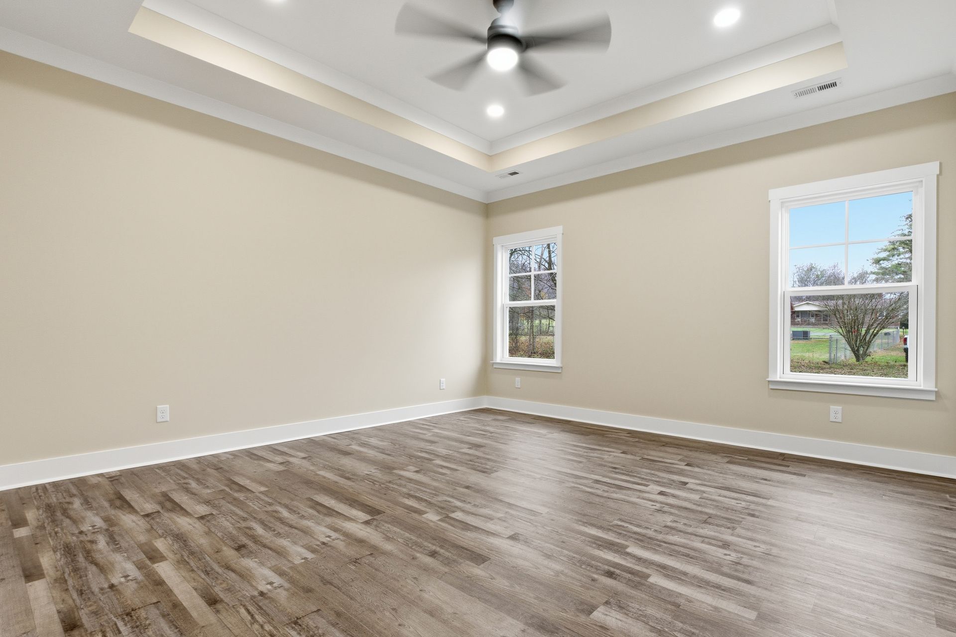 Empty bedroom with wood-look flooring, beige walls, two windows, and a ceiling fan.