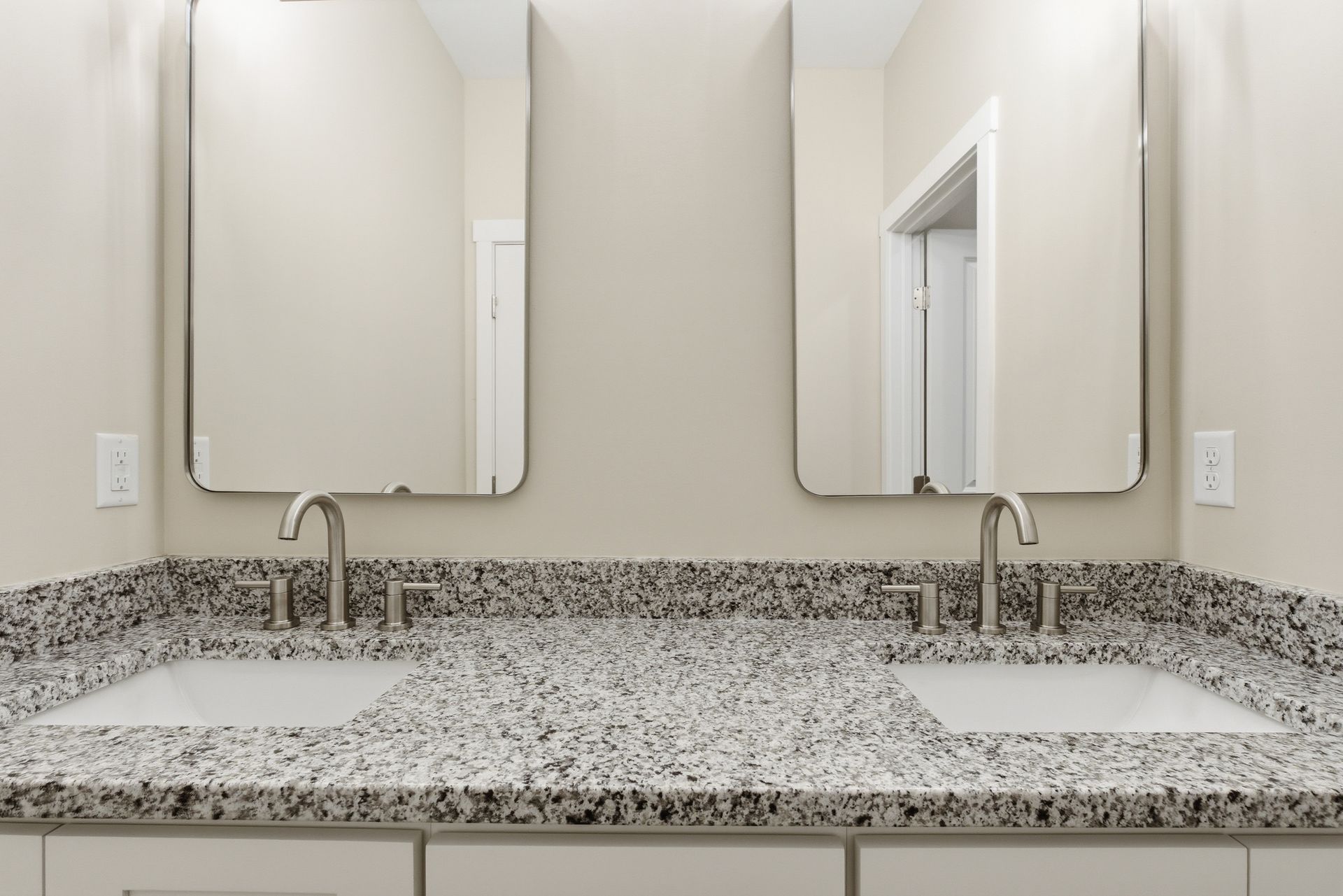 Bathroom with dual sinks, speckled granite countertop, two square mirrors, and a neutral wall.