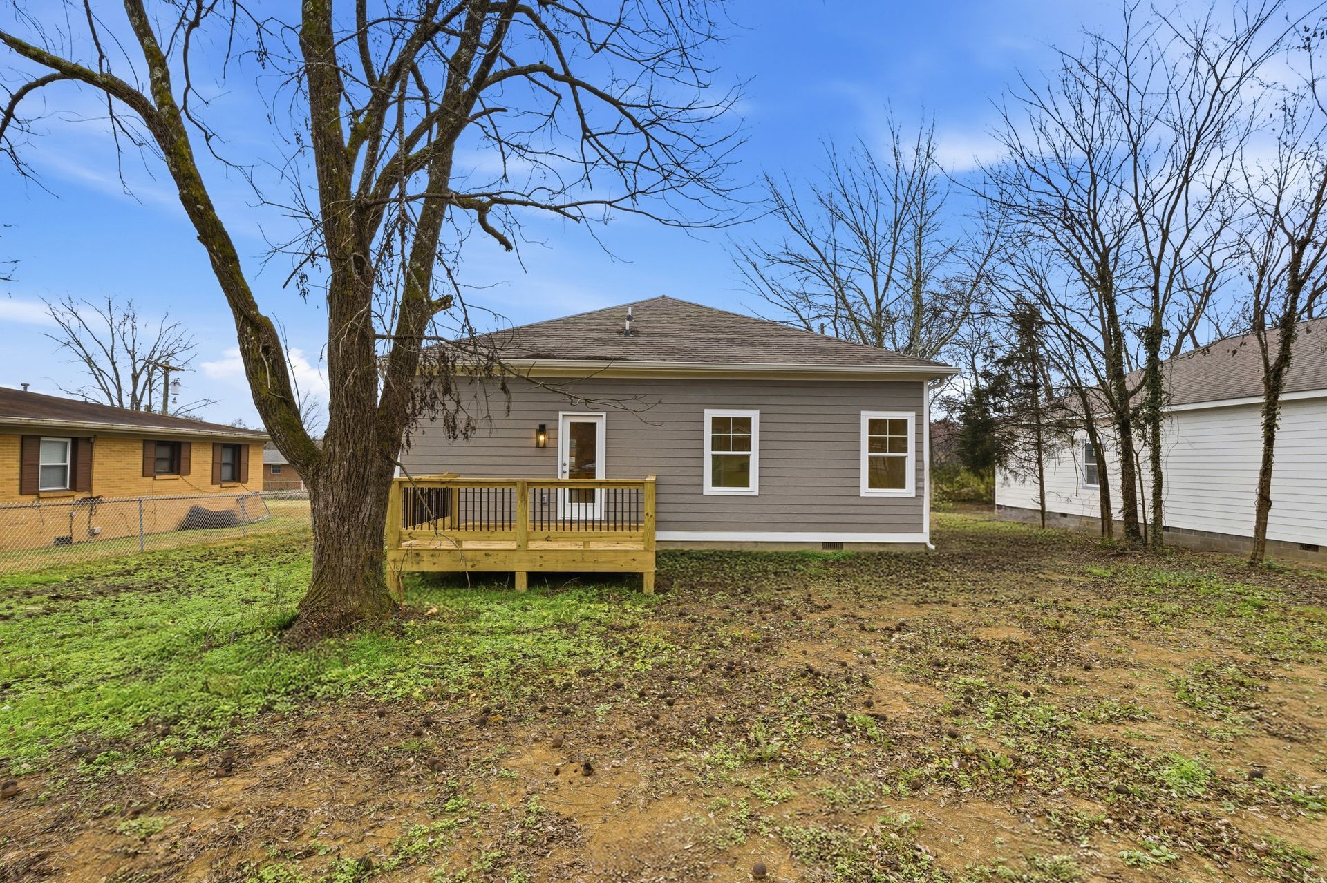 Back view of a small, gray house with a wooden deck and bare trees against a blue sky.