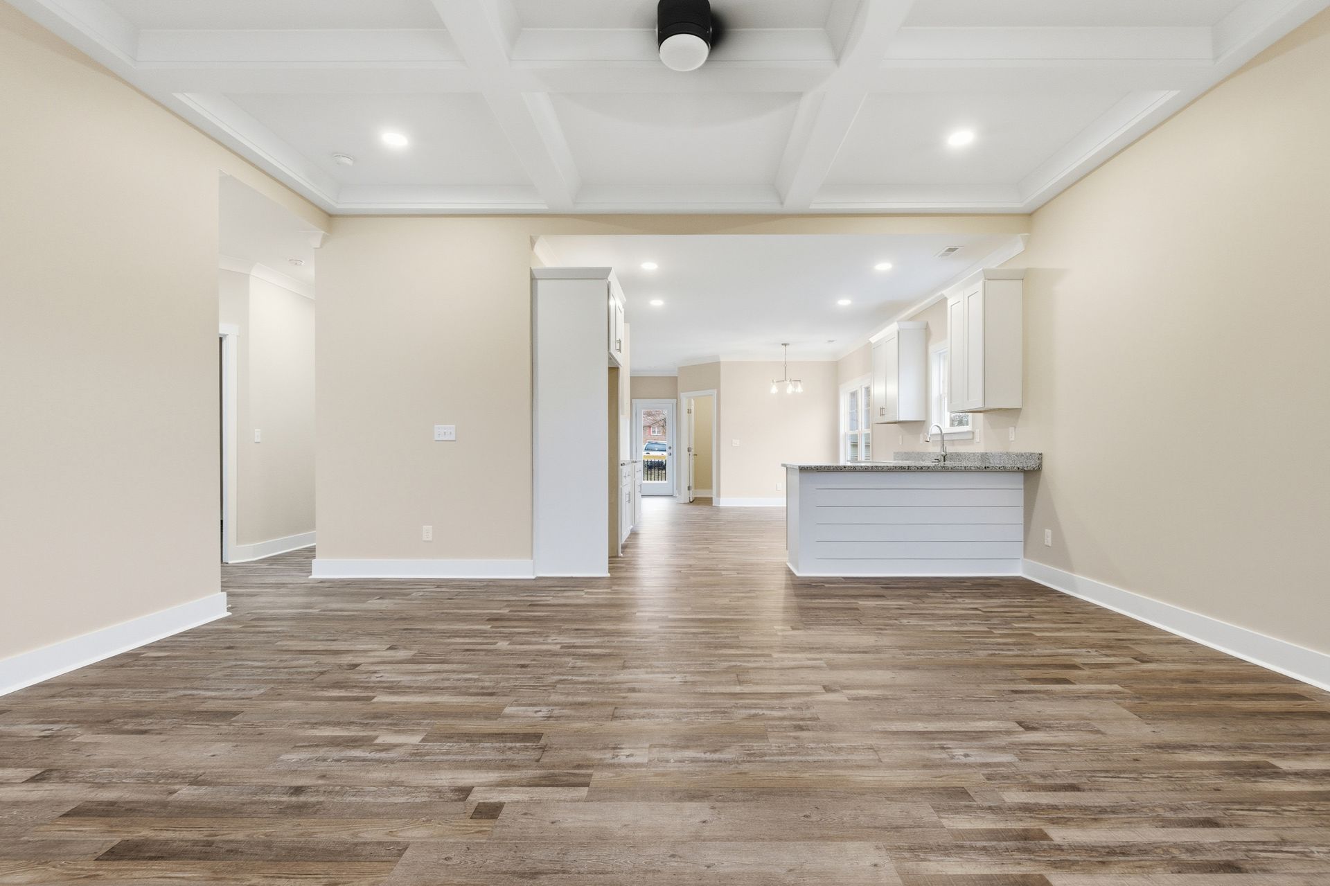 Empty living space with wood-look flooring, neutral walls, and a view into the kitchen area.
