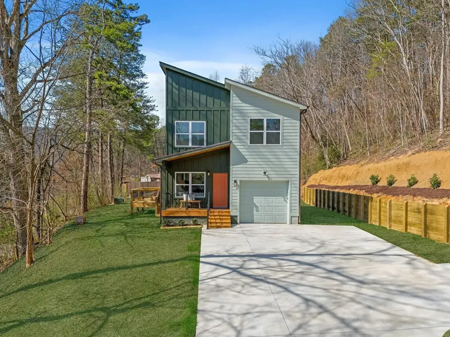 A house with a green roof and a garage is sitting on top of a hill surrounded by trees.