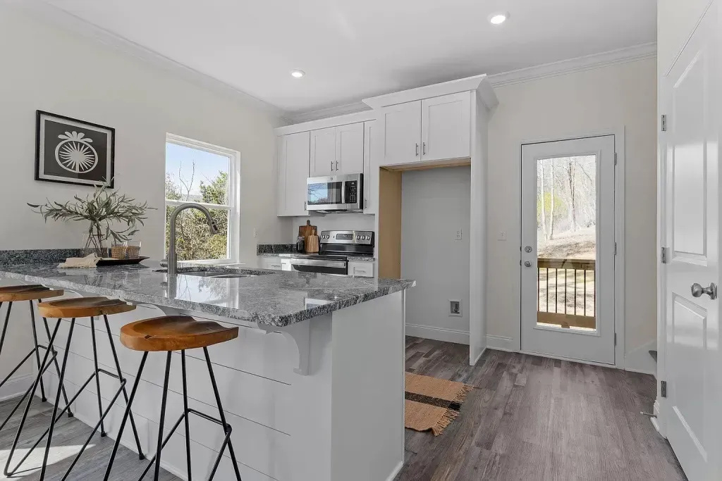 A kitchen with white cabinets , granite counter tops , stools and a window.
