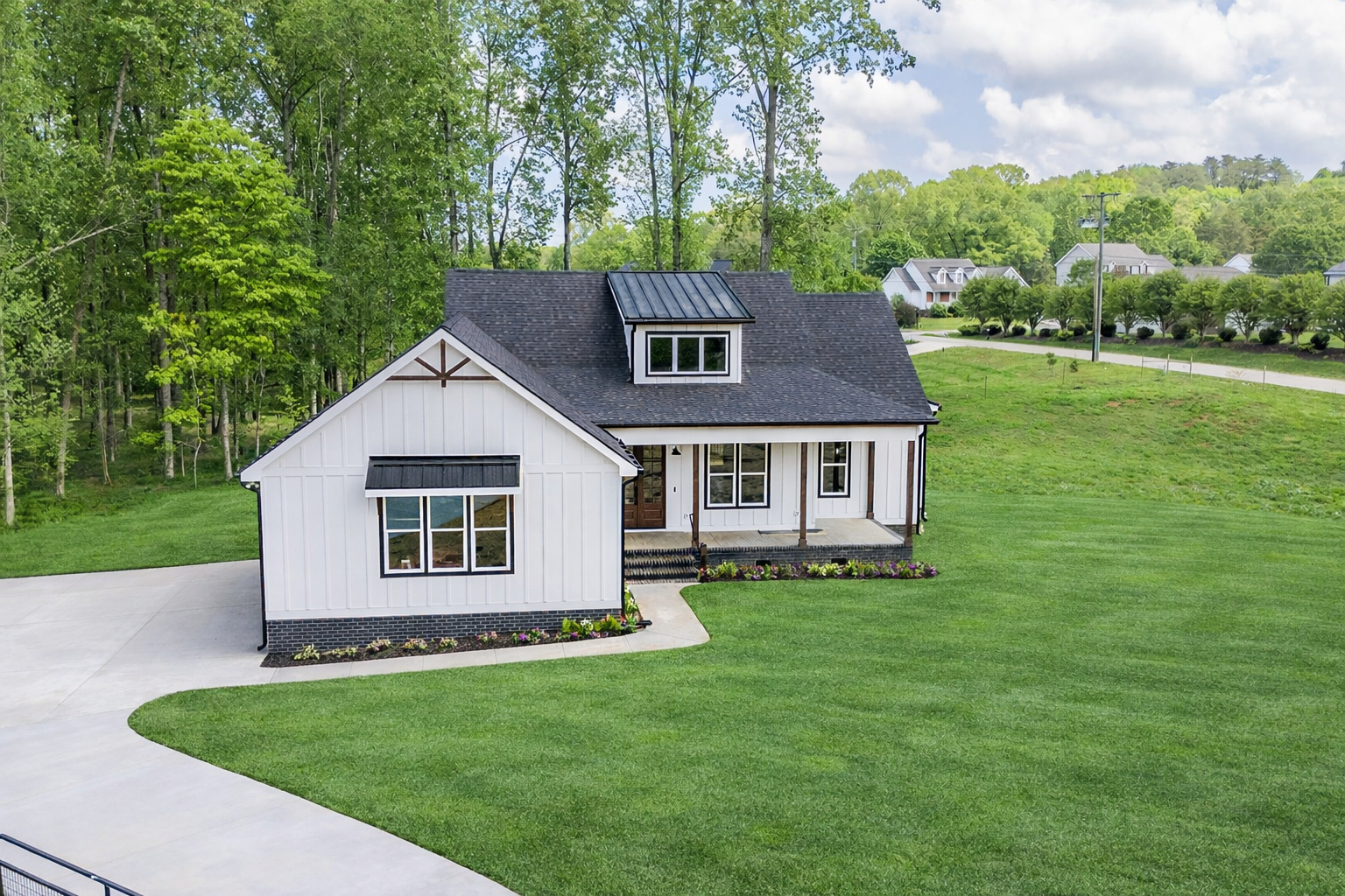 White house with black roof, porch, and concrete driveway on a grassy lot with trees.
