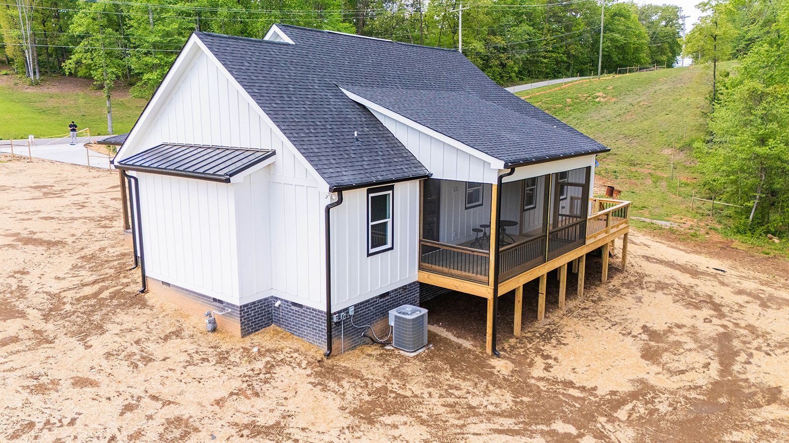 White house with black roof and screened porch on a brown dirt lot, with a green hill in the background.