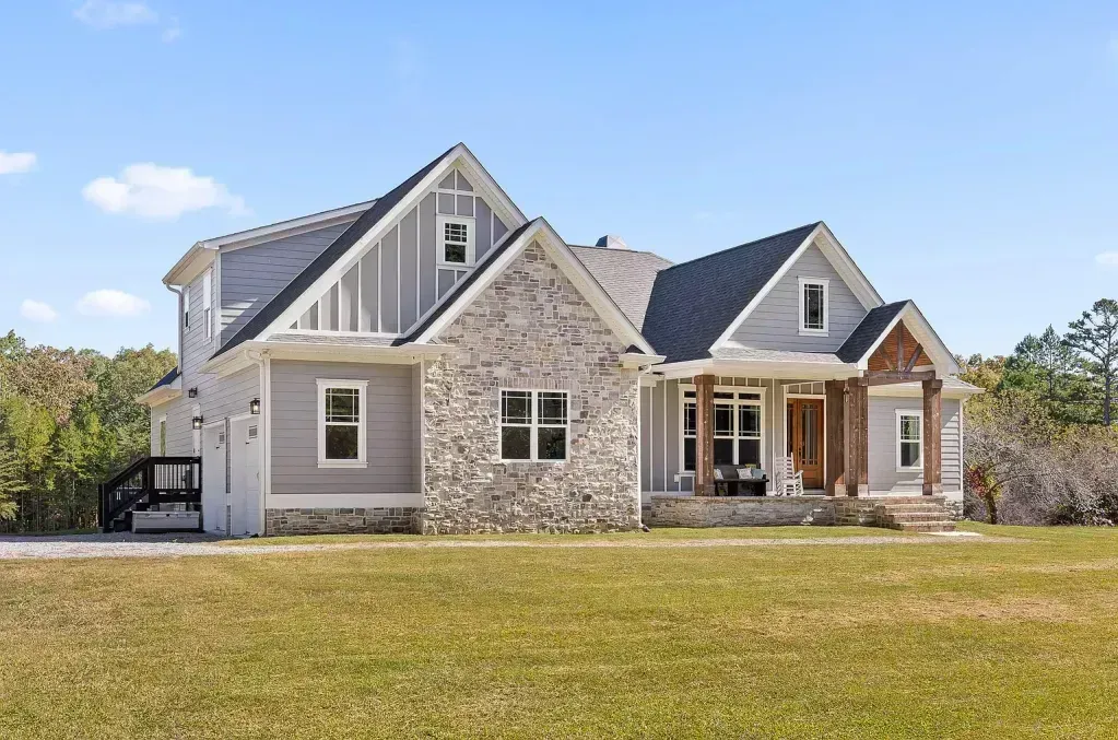 Gray and stone Craftsman-style house with porch, lawn, and trees in background under a blue sky.