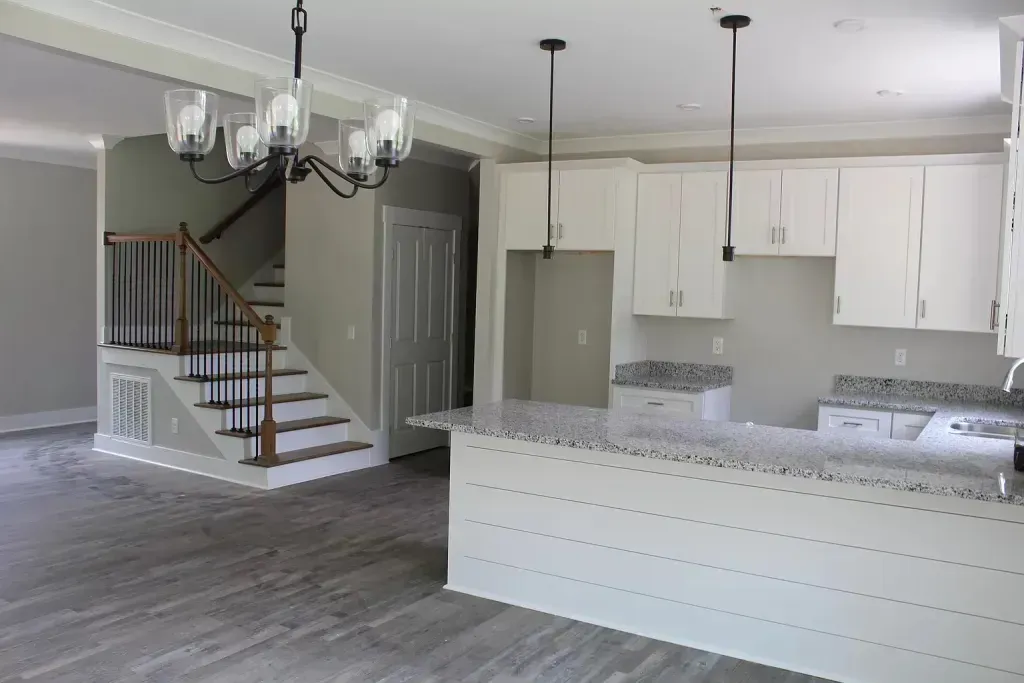 An empty kitchen with white cabinets , granite counter tops , a staircase and a chandelier.