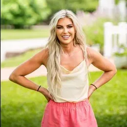 A woman in a white tank top and pink skirt is standing in the grass with her hands on her hips.