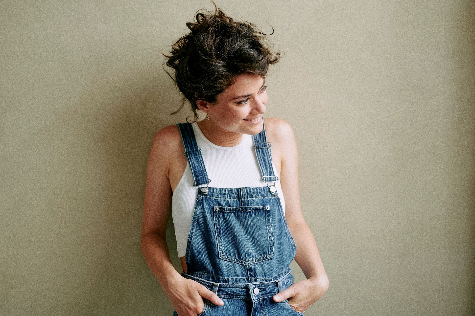 Eine Frau in einem Overall und einem weißen Tank Top steht vor einer Wand.