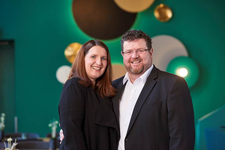 Ein Mann und eine Frau posieren für ein Foto vor einer grünen Wand.