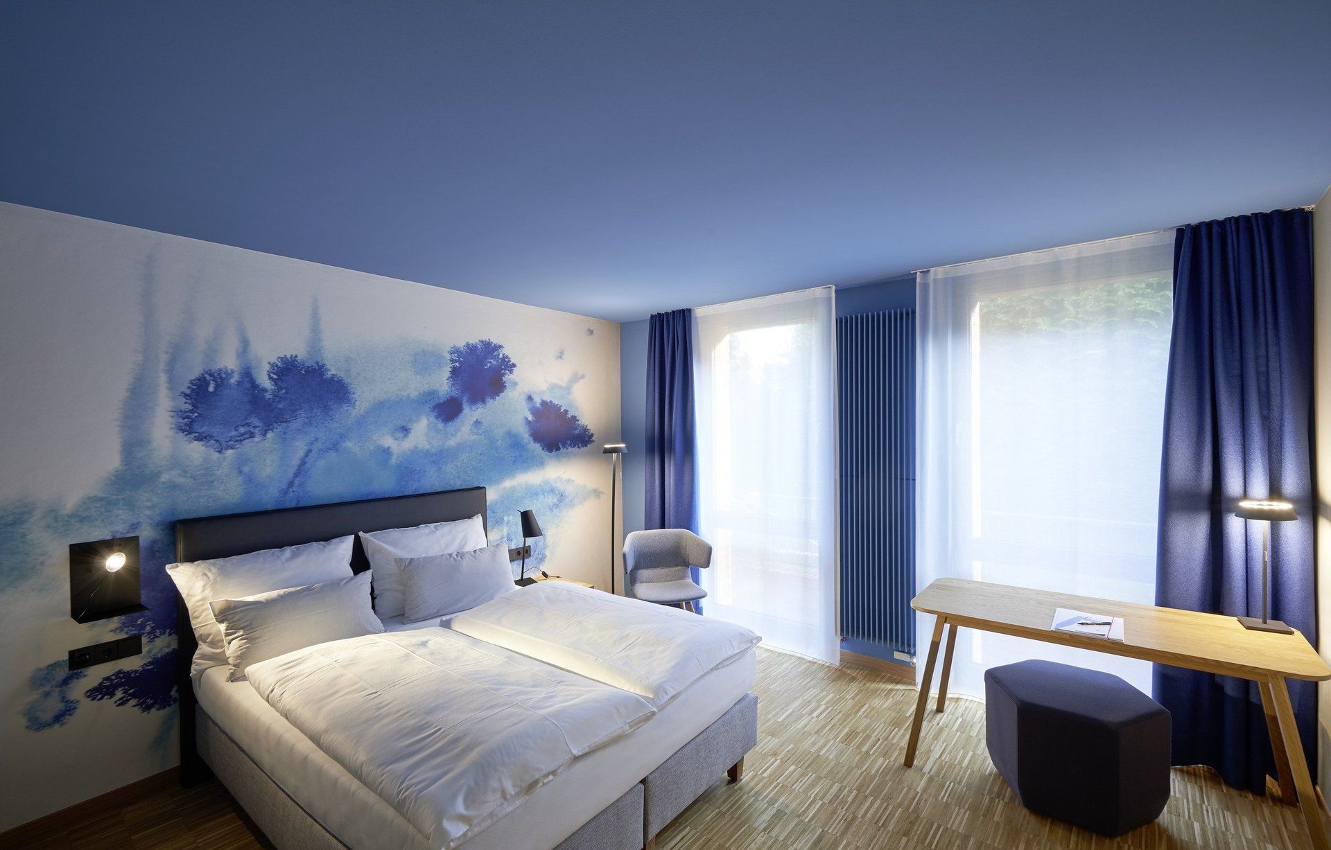 Ein Hotelzimmer mit Bett, Schreibtisch, Stuhl und blauen Vorhängen.