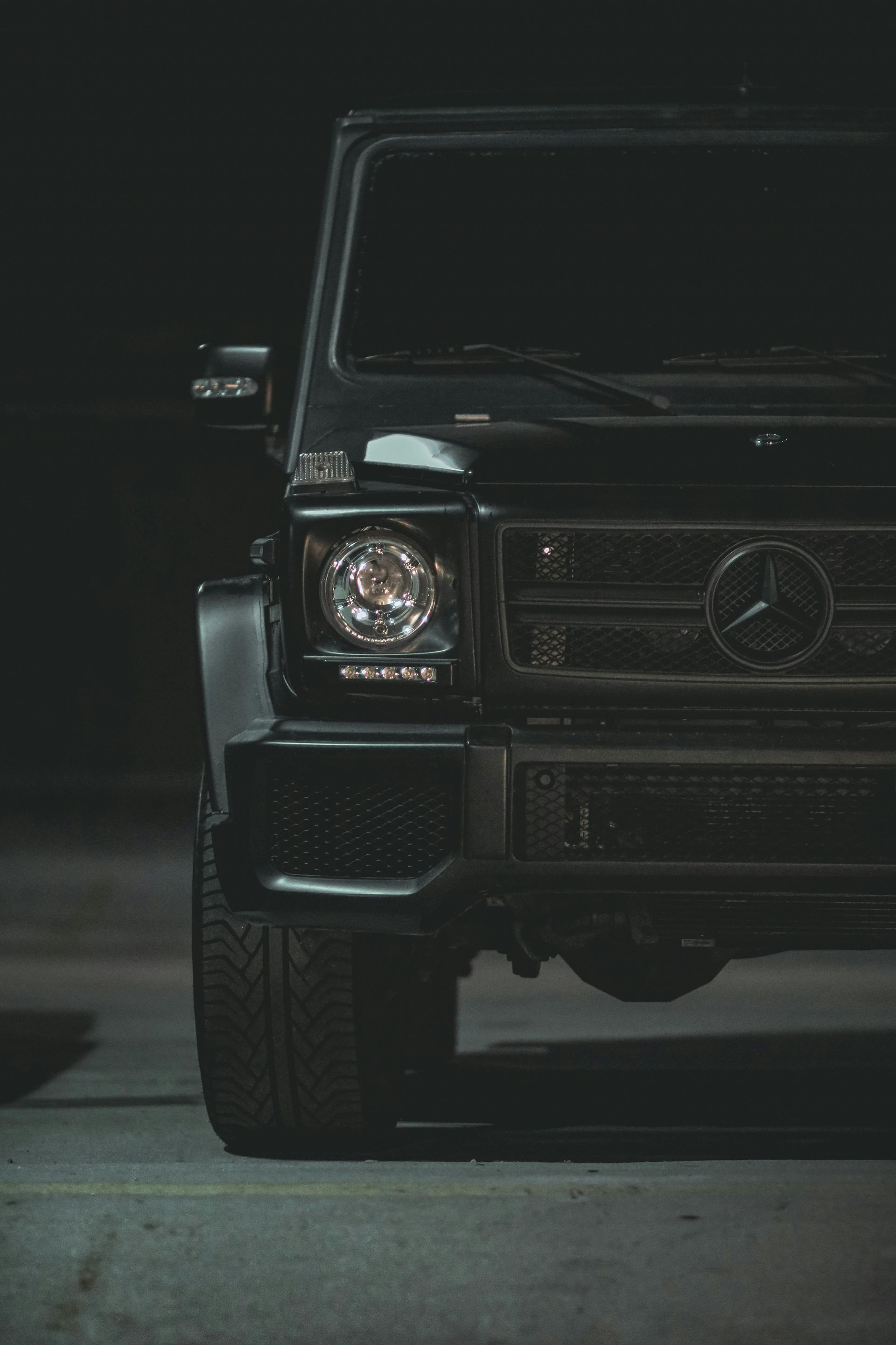 Black Mercedes-Benz G-Wagon SUV. Front view with headlights and grill visible. Dark setting.