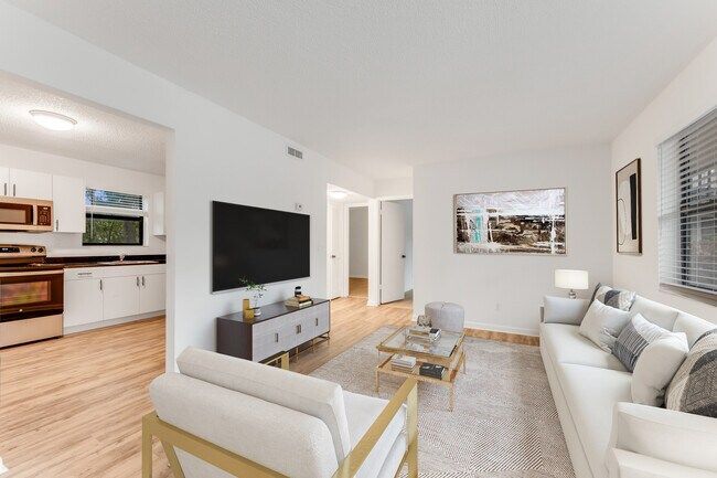 Living room with white sofa, gold chair, TV, and glass coffee table. Kitchen visible in background.