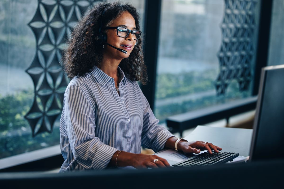 Woman with headset types on keyboard at a desk, looking at a computer screen. Modern office setting.