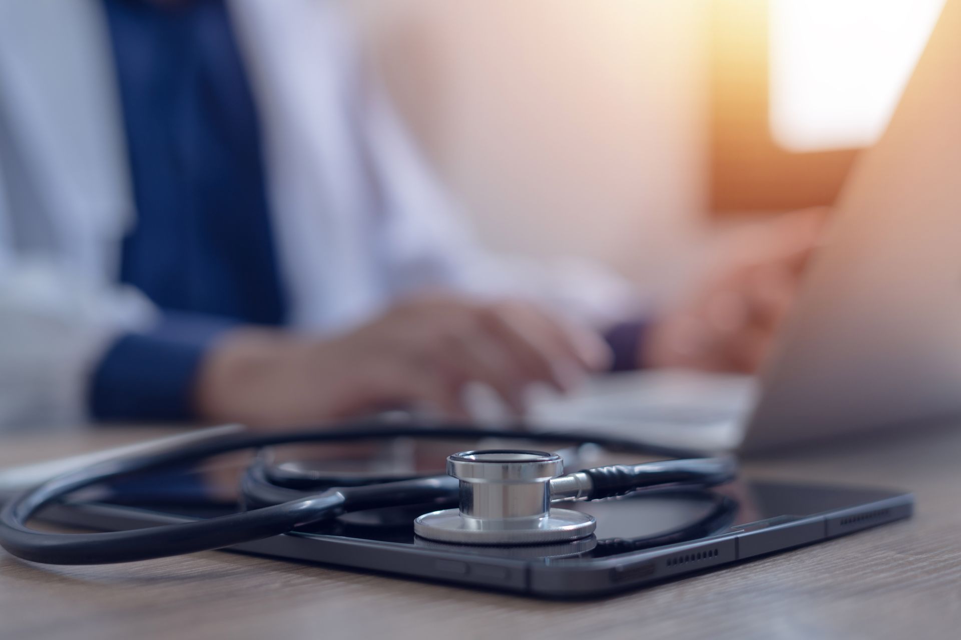 A stethoscope is sitting on top of a tablet next to a doctor using a laptop.