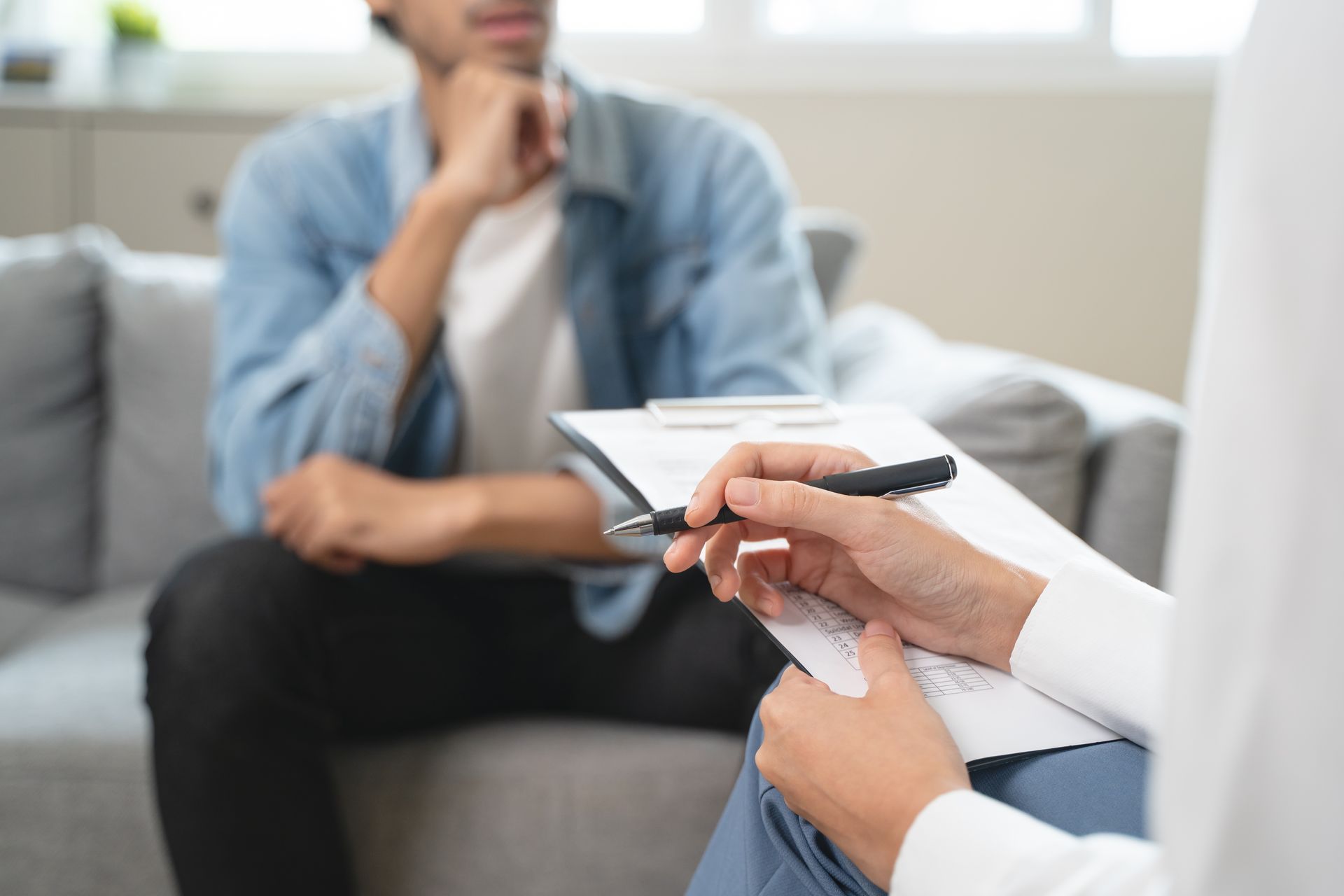 A man is sitting on a couch talking to a woman who is holding a clipboard.