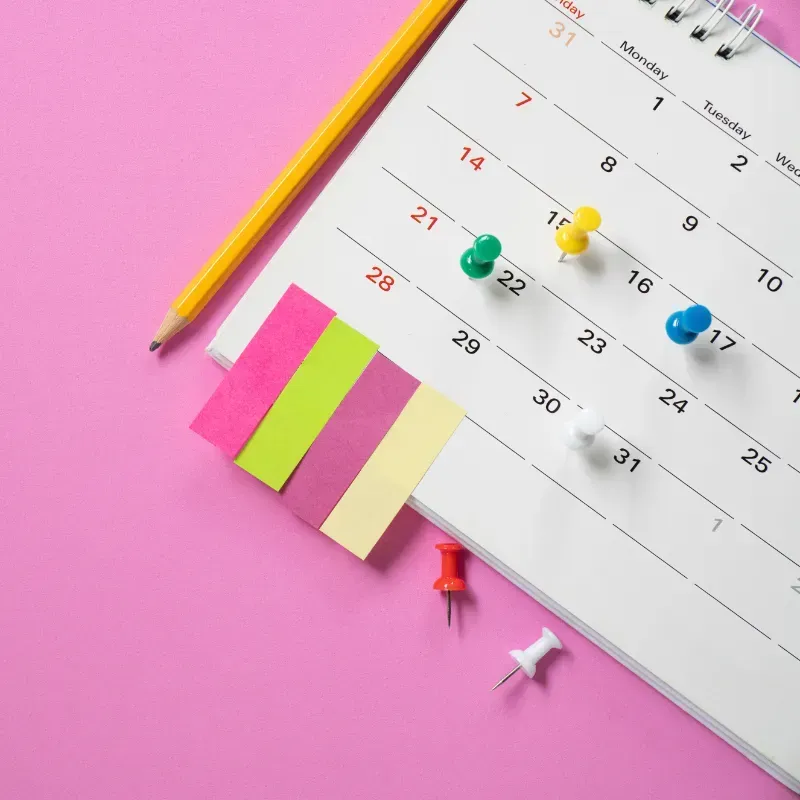 A calendar with sticky notes , pins and a pencil on a pink background.