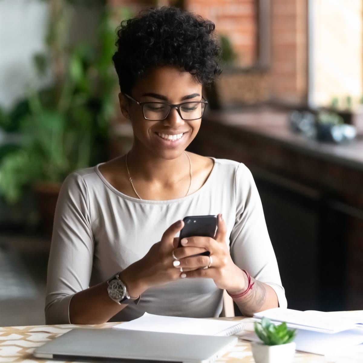 Woman with glasses smiles while looking at a phone, seated at a desk with laptop, papers, and a plant.