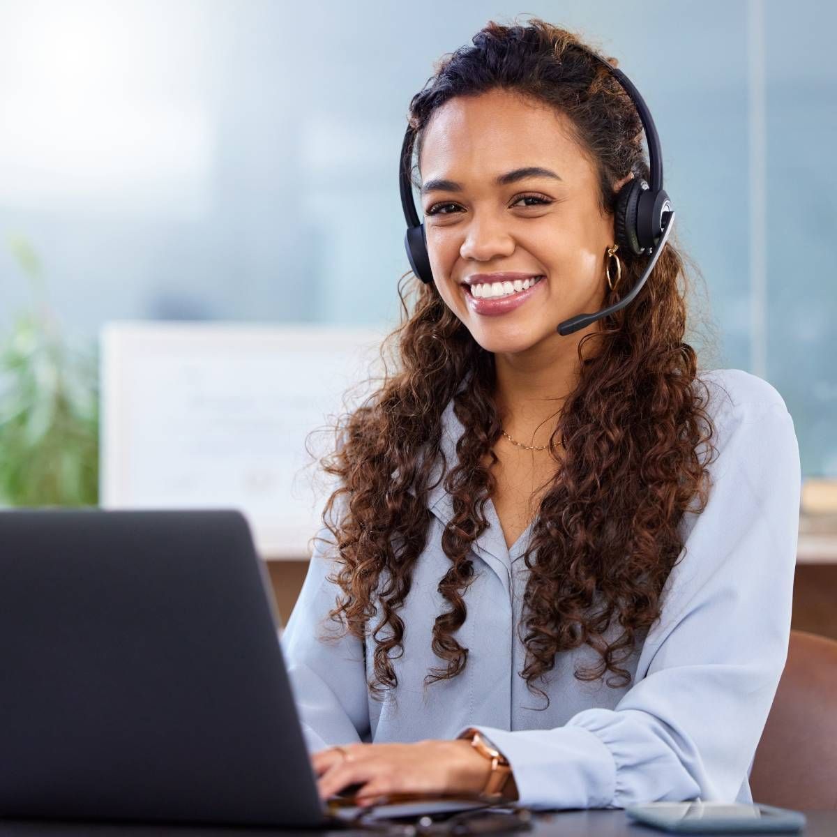 Woman with curly hair wearing headset, smiling at laptop, in office.