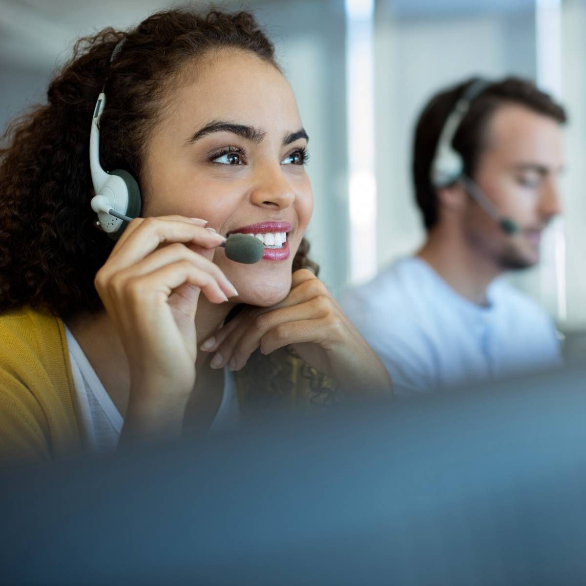 Woman with headset smiling, supporting a customer service role. Another person in background.
