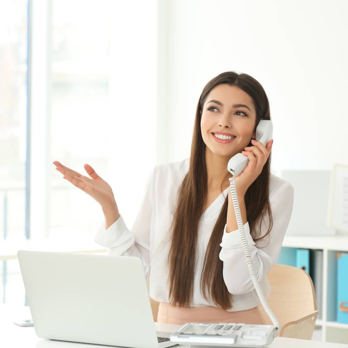 Woman smiling while talking on a landline phone at her desk, gesturing with her left hand.