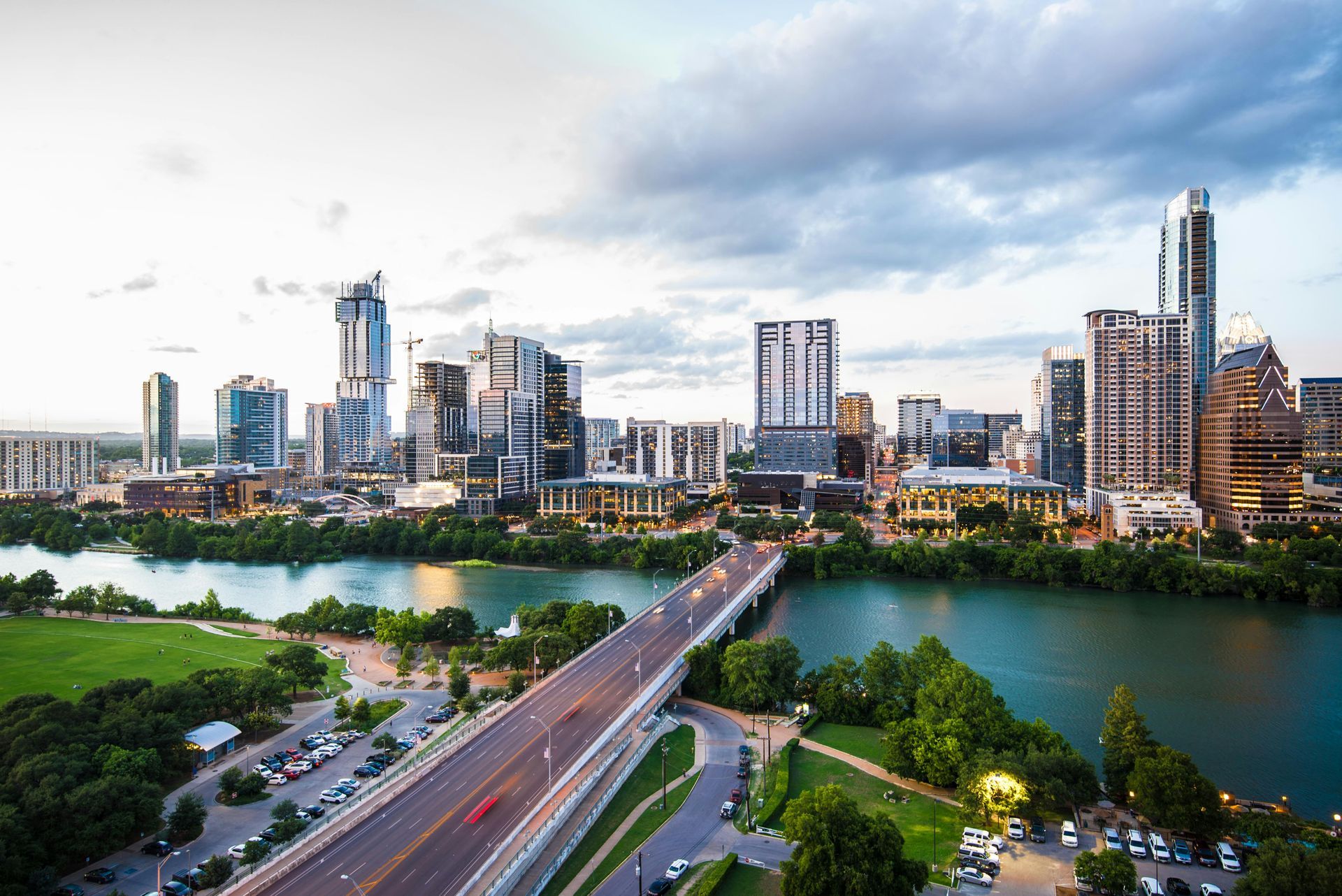 Austin, Texas skyline with bridge over a river, illuminated buildings, and green space, at dusk.