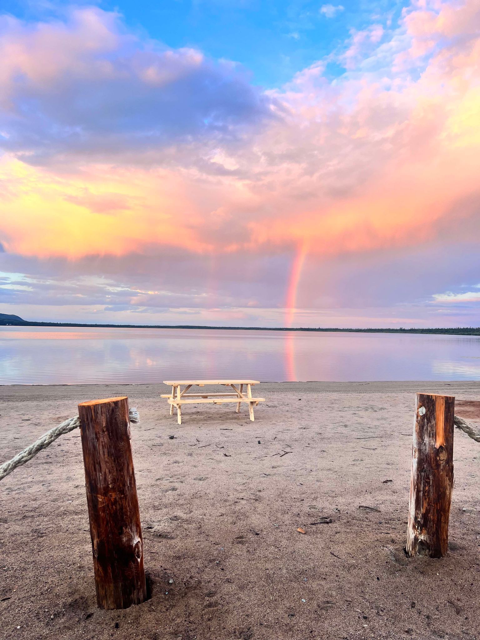 A picnic table on a beach with a rainbow in the sky.