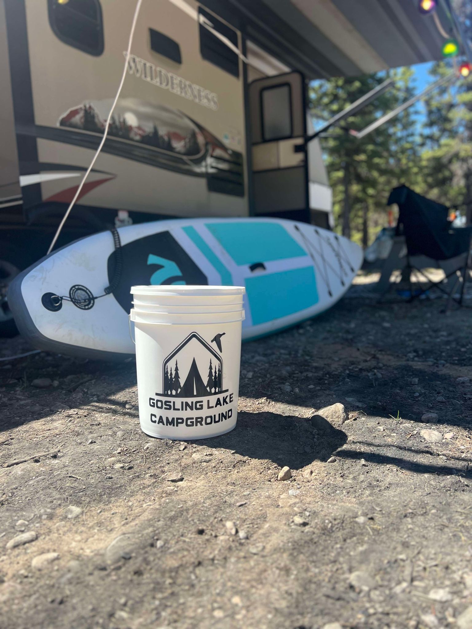 A bucket is sitting on the ground next to a paddle board and a rv.