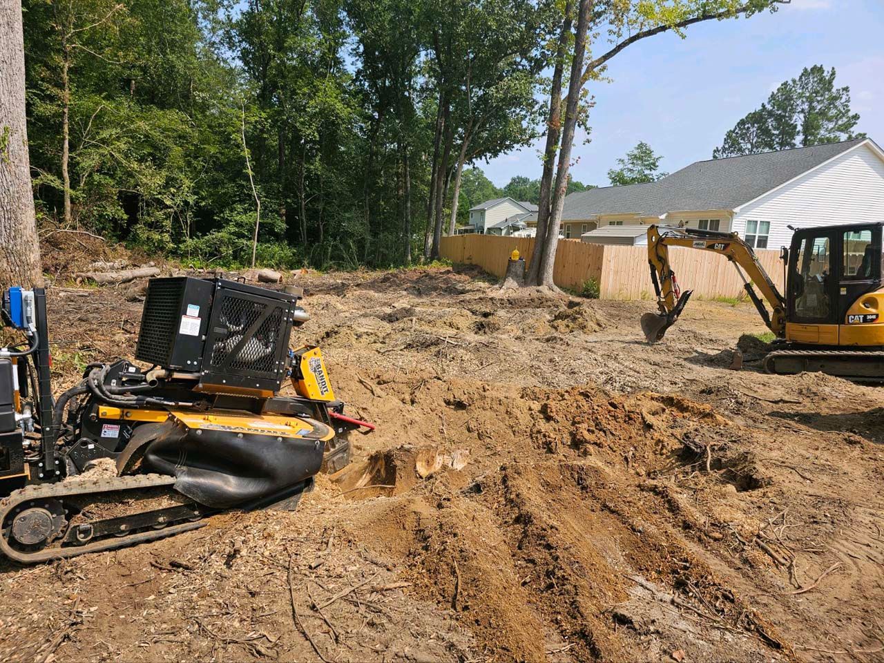A bulldozer and a stump grinder are in a dirt field in front of a house.