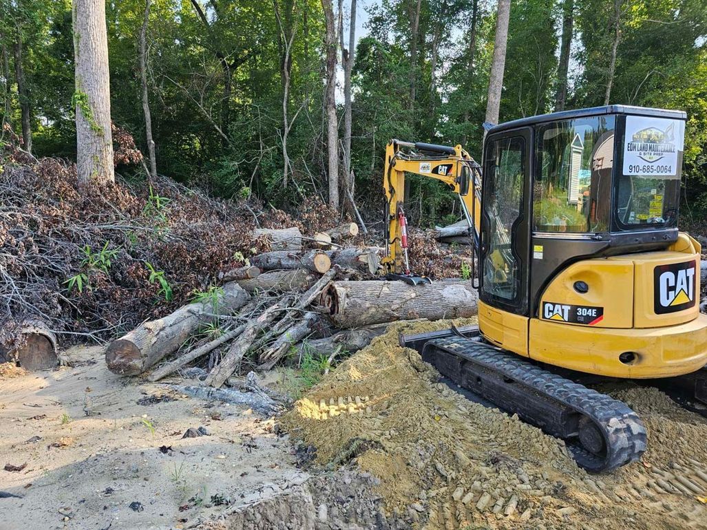 A yellow cat excavator is sitting in the dirt next to a pile of logs.