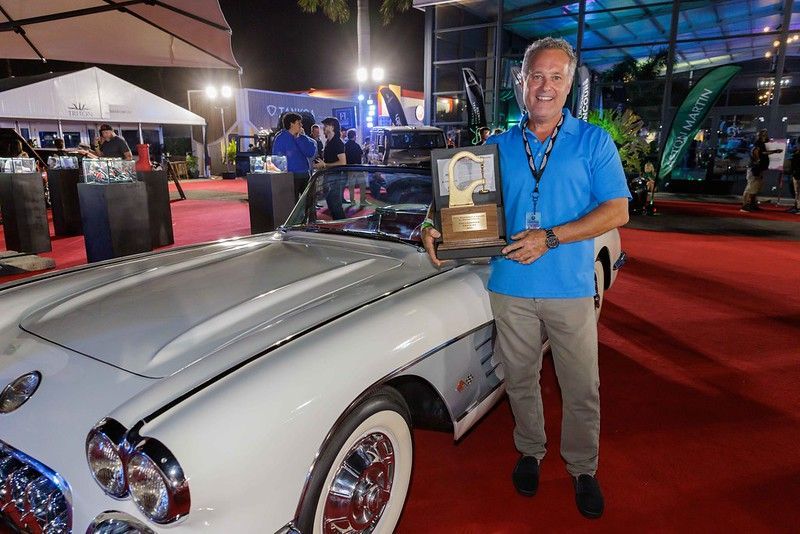 A man is standing in front of a white car holding a trophy.