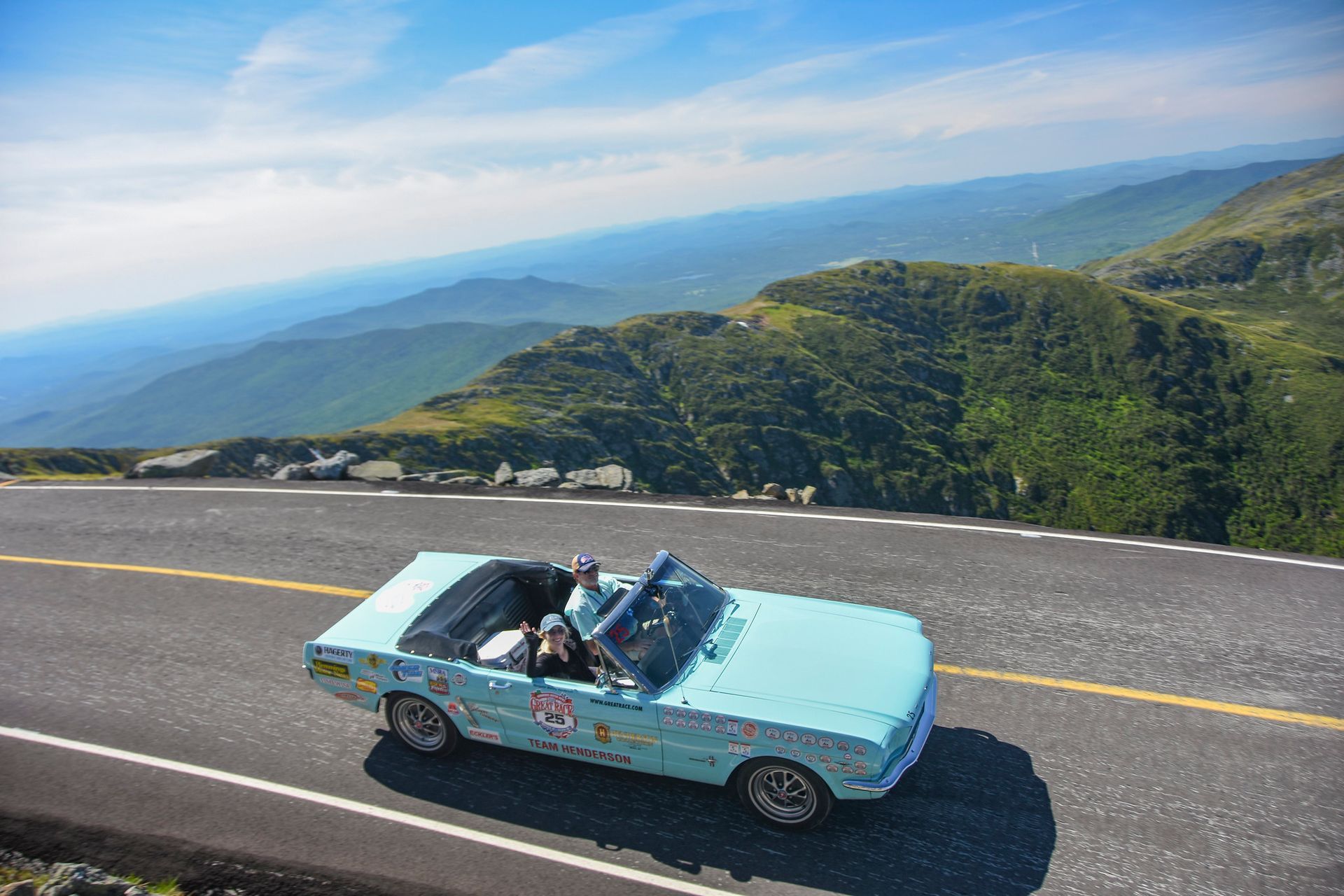 A blue convertible car is driving down a mountain road.
