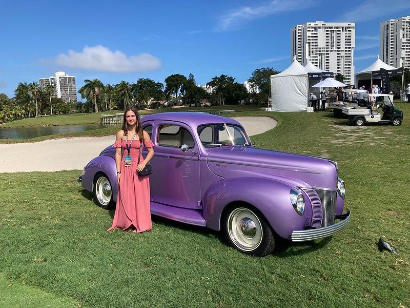 a classic purple car on a green field with a woman in a dress nearby