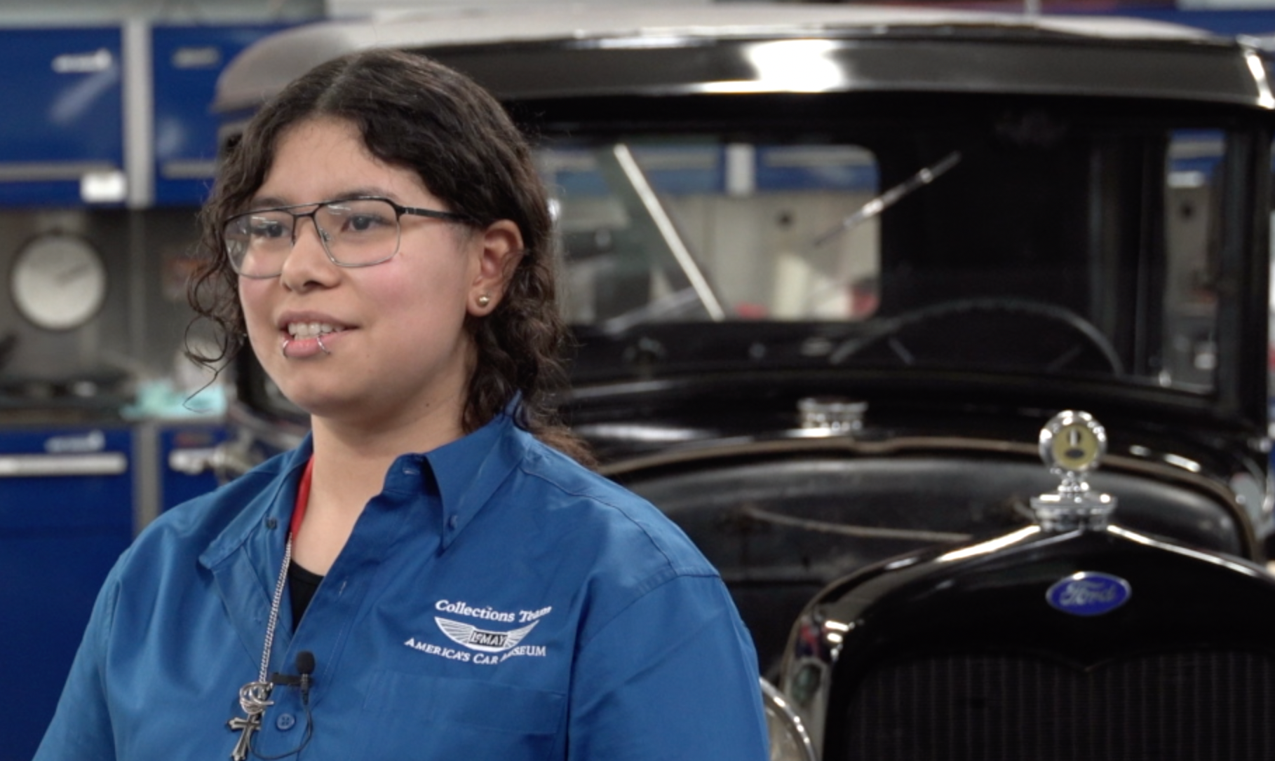 Woman in blue shirt speaks in a garage next to a black vintage Ford car.