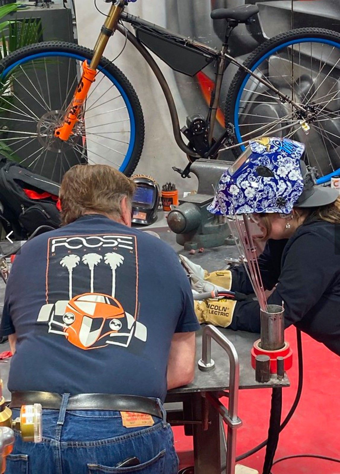 Two people welding at a table, a mountain bike behind them. One wears a blue t-shirt with a car graphic.