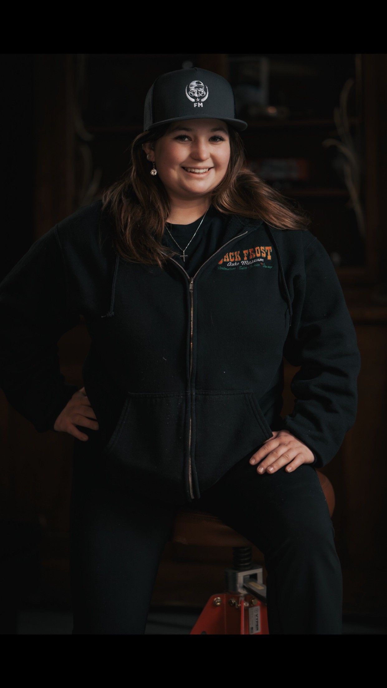 Woman in black jacket and cap with logo, smiling, hand on hip, seated.