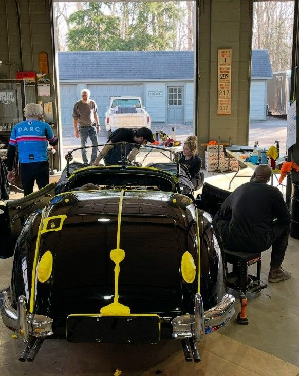 A black classic car being worked on in a garage. People are gathered around the car.